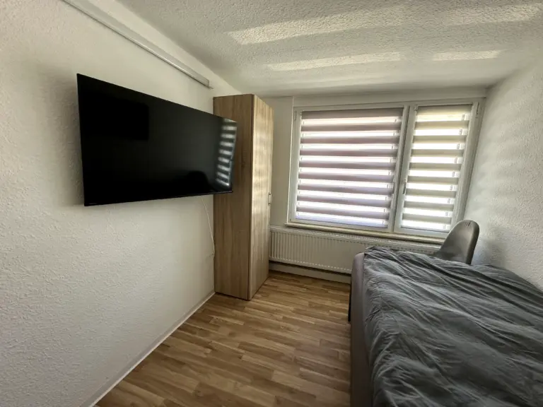 Ein Einzelbettzimmer mit großem Flachbildfernseher, Kleiderschrank und Fenster mit Plissees in Monteurunterkunft bei Jena