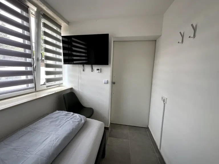 Kompaktes Einzelzimmer mit Bett, Fernseher, Fensterrollos und Stuhl – Monteurzimmer bei Jena